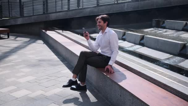 Mensajería de texto alegre hombre de negocios en el teléfono móvil sentado en el banco — Vídeos de Stock