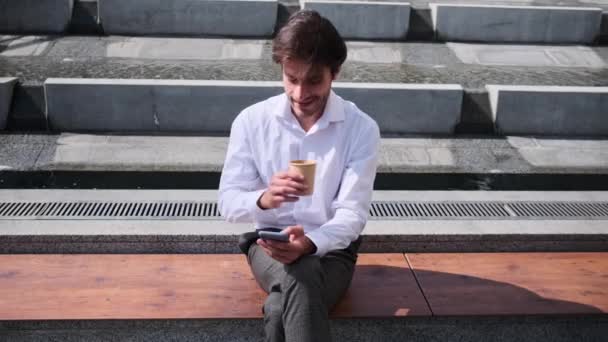 Mladý muž pije kávu a pomocí mobilního telefonu sedí na lavičce — Stock video
