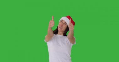 Noel Baba şapkalı heyecanlı kadın yeşil arka planda başparmak hareketi yapıyor.