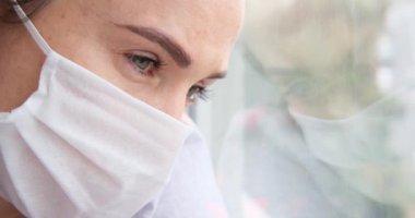 Pencereye yaslanmış, koruyucu yüz maskesi takan üzgün, yalnız bir kadın.