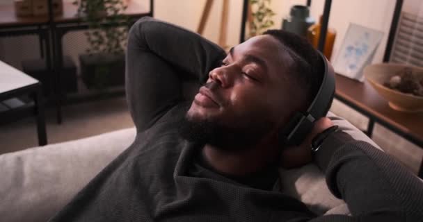 Homem ouvindo música em fones de ouvido enquanto se inclina no sofá — Vídeo de Stock
