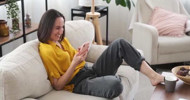 Mujer usando teléfono móvil tumbado en el sofá en casa — Vídeo de stock