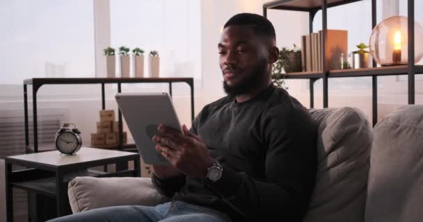 ชายชาวแอฟริกันอเมริกันยิ้มขณะนั่งบนโซฟาและใช้แท็บเล็ตดิจิตอล — วีดีโอสต็อก