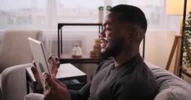 Dijital tablette kanepeye oturup video konferans veren mutlu genç adam.