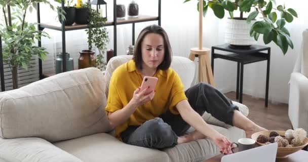 Γυναίκα που χρησιμοποιεί το κινητό τηλέφωνο ξαπλωμένη στον καναπέ στο σπίτι — Αρχείο Βίντεο