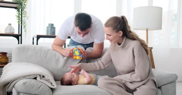 Parents playing with rattle over baby son lying on sofa — ストック動画