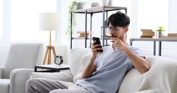 Чоловік використовує мобільний телефон, сидячи на дивані вдома — стокове відео