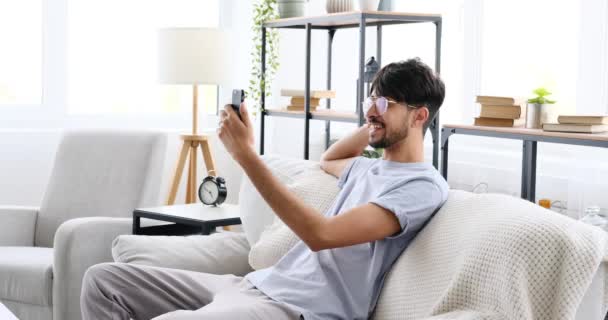 Man video chatting using mobile phone sitting on sofa at home — Vídeo de Stock