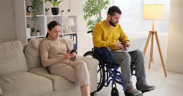 Homem com deficiência trabalhando em laptop e esposa usando telefone celular — Vídeo de Stock