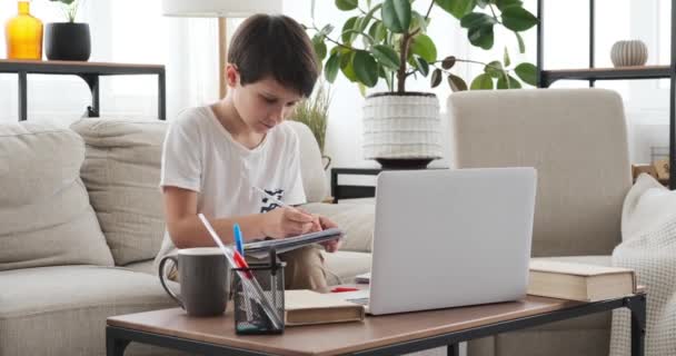 Chłopiec za pomocą laptopa i pisania w książce w domu — Wideo stockowe