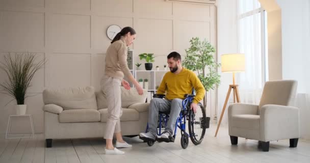 Wife helping disabled husband to sit on sofa from wheelchair — Vídeo de Stock