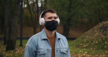 Koruyucu yüz maskesi takan bir adam parkta kulaklıkla müzik dinliyor.