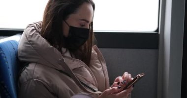 Otobüste cep telefonu kullanan koruyucu maskeli bir kadın.