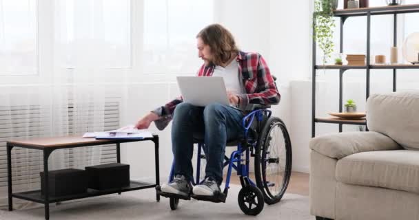 Homem com deficiência sentado em cadeira de rodas trabalhando no laptop e analisando documentos — Vídeo de Stock