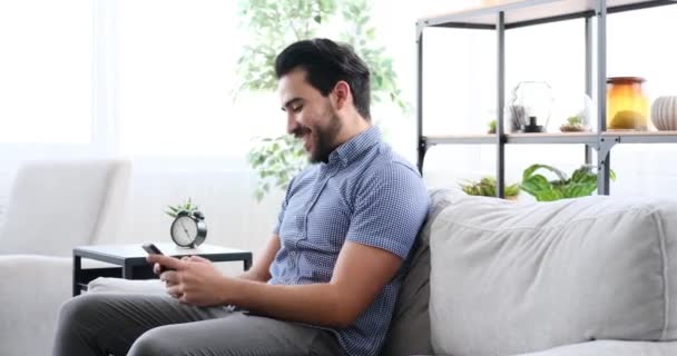 Молодий чоловік використовує мобільний телефон, сидячи на дивані — стокове відео