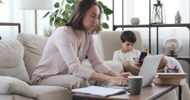 息子が自宅で携帯電話を使っている間にラップトップで働く母親 — ストック動画