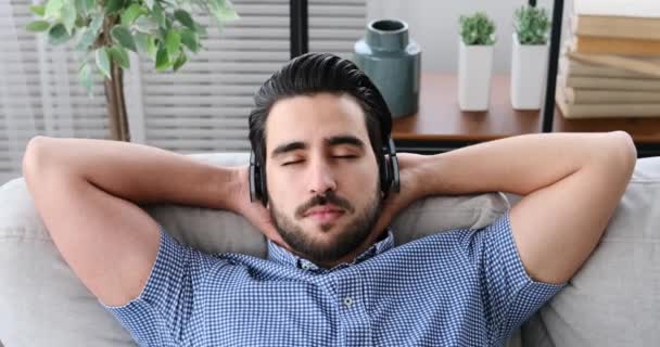 Jovem ouvindo música em fones de ouvido enquanto relaxa no sofá — Vídeo de Stock