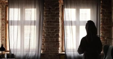 Bornozlu kadın perdeyi açıyor ve pencereden dışarı bakıyor.