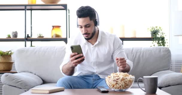 Молодий чоловік весело слухає музику на навушниках і використовує мобільний телефон під час їжі попкорн — стокове відео