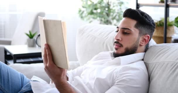 Hombre feliz leyendo libro mientras se relaja en el sofá — Vídeo de stock