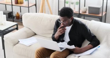 Genç Afro-Amerikalı iş adamı dizüstü bilgisayar kullanıyor ve kanepede belgeleri analiz ediyor.