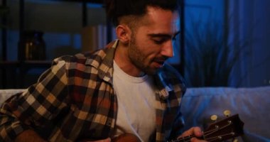 Genç adam evde gitar çalıp şarkı söylüyor.