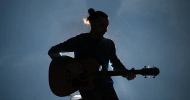 Gitar çalan genç adam taburede oturuyor.