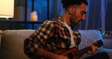 Genç adam kanepede oturup gitar çalıp şarkı söylüyor.