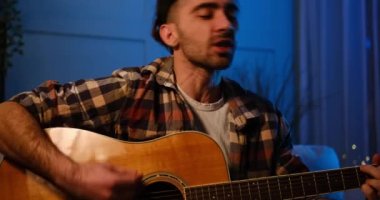 Genç adam evde şarkı söylüyor ve gitar çalıyor.
