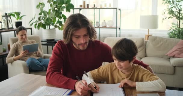 Padre ayudando a su hijo a hacer los deberes y madre usando tableta digital — Vídeo de stock