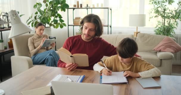 Отец помогает сыну делать домашнее задание из учебника и матери с помощью цифрового планшета — стоковое видео