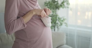 Evde hamile karnında bebek ayakkabıları taşıyan neşeli bir kadın.