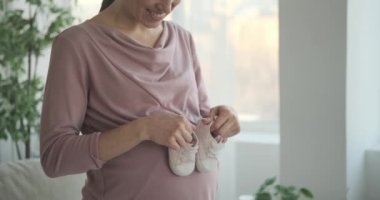 Evde hamile karnında bebek ayakkabıları taşıyan neşeli bir kadın.