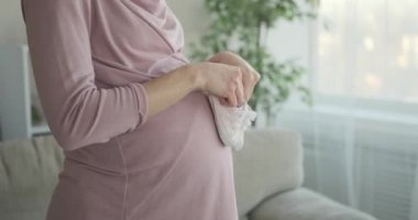 Evde hamile karnında bebek ayakkabıları taşıyan neşeli bir kadın.