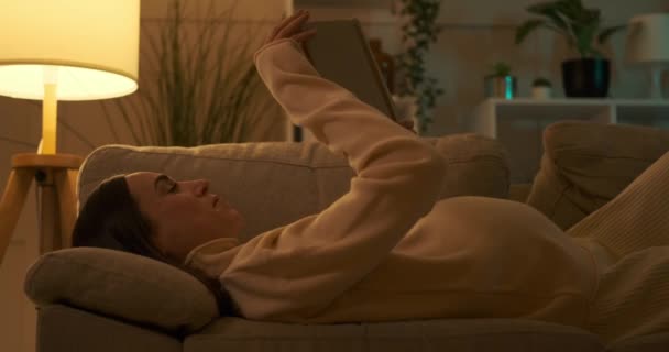 Pregnant woman reading book on sofa at night — Stock Video