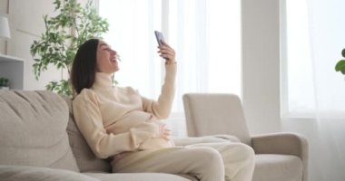 Hamile kadın cep telefonuyla evde görüntülü konuşma yapıyor.