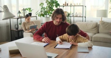 Baba oğluna masa başında ödev yaparken yardım ediyor ve anne dijital tablet kullanıyor.