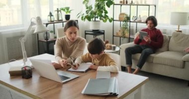Anne oğlunun ödevlerine yardım ediyor ve baba dijital tablet kullanıyor.