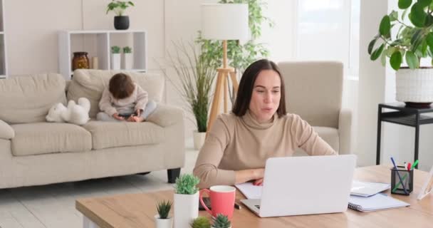 Mulher vídeo conversando no laptop em casa, enquanto a filha está usando telefone celular no sofá — Vídeo de Stock