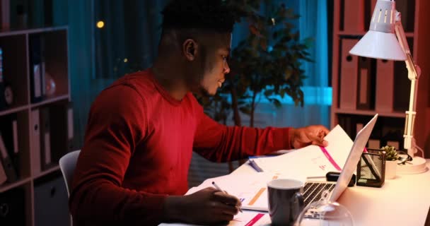 Afroamerykanin biznesmen korzystający z laptopa i wykonujący papierkową robotę do późna — Wideo stockowe