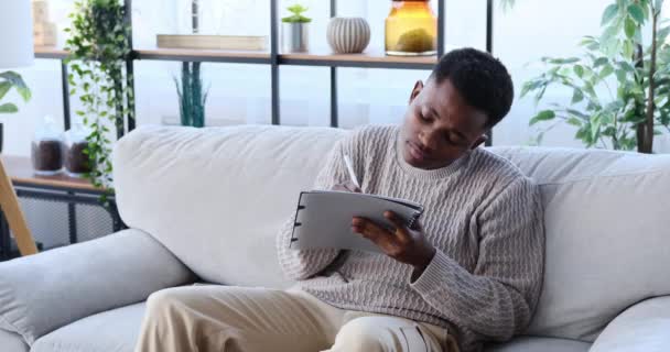 Νεαρός άντρας γράφει για να κάνει λίστα στο σημειωματάριο κάθεται στον καναπέ — Αρχείο Βίντεο