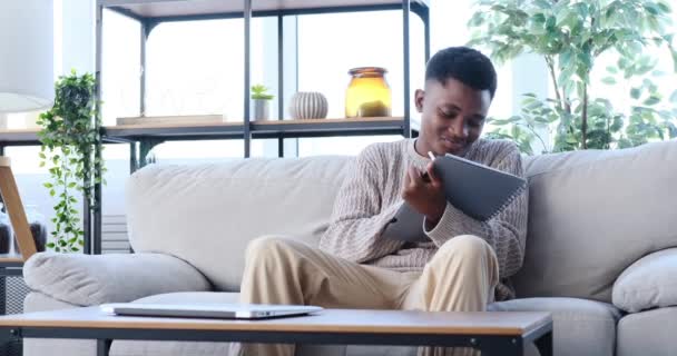Afroamerikansk man skriver för att göra lista i anteckningsblock sitter på soffan — Stockvideo