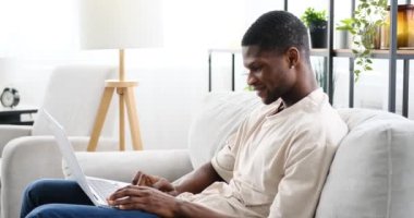 Evdeki kanepede otururken dizüstü bilgisayarda çalışan Afrikalı Amerikalı bir adam.