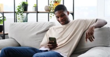 Afro-Amerikalı adam evdeki koltukta otururken cep telefonuyla sohbet ediyor.