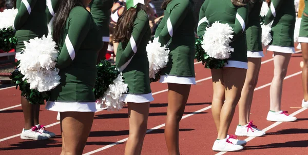 High school cheerleaders standing on a track holding their white pom poms behind their backs during a football game.