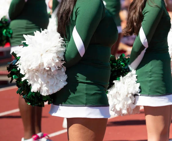 Close up of high school cheerleaders in green uniforms holding white and green pom poms behind their backs.