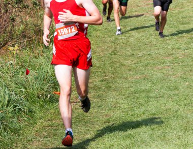 Bir liseli çocuk, 5 km çapındaki bir kır koşusunda tepeden aşağı doğru kırmızı üniformalı bir çim tarlasında koşuyor..