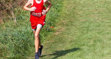 5K 'lık bir kros yarışında yarışan liseli bir çocuk tepeden aşağı çim tarlasında kırmızı üniformalı koşuyor..