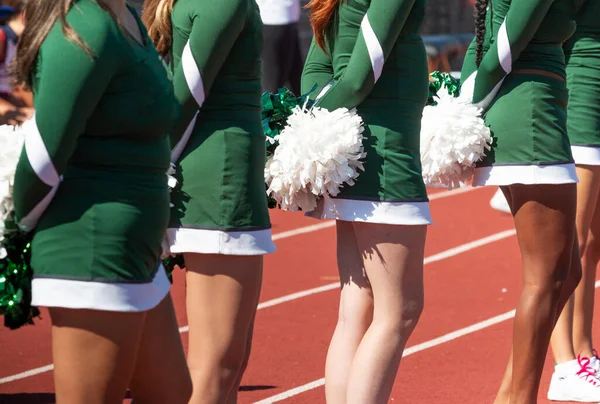 Close up of high school cheerleaders standing on a track watching the football game holding their pom poms behind their backs.