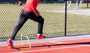 Bir liseli koşucu lisede kırmızı pistte 60 santimlik sarı bir engeli aşarak hız talimi yapıyor..